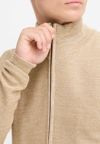 Suéter beige de cremallera con cuello alto, patrón de punto texturizado y mangas largas ajustadas. La cremallera cuenta con un tirador metálico.