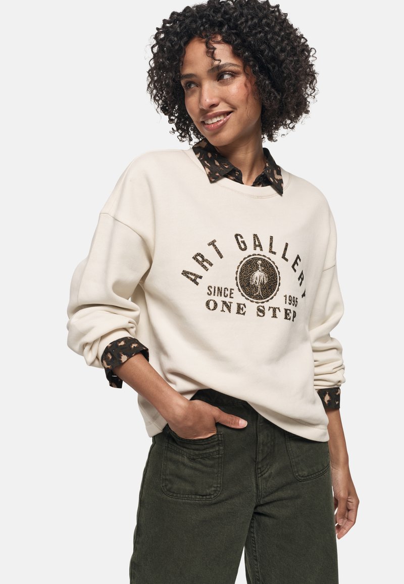 Kremowy sweatshirt z okrągłym graficznym logo, z ciemno-brązowym napisem. Pod spód wystaje czarna koszula z delikatnym wzorem.