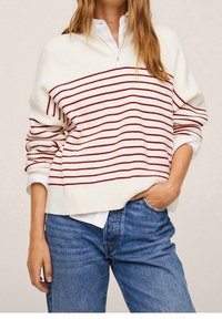 Donna che indossa un maglione bianco con strisce orizzontali rosse sopra una camicia bianca e dei jeans blu, con una mano nella tasca e l'altra rilassata.