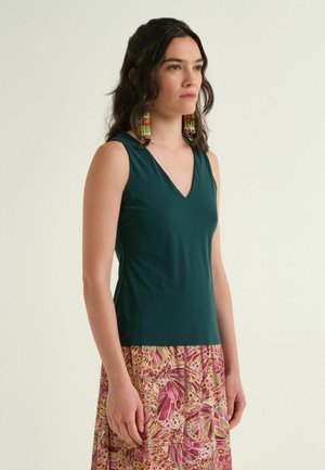 Donna che indossa un top verde senza maniche con scollo a V, gonna colorata a fantasia e grandi orecchini con perline, in piedi davanti a uno sfondo chiaro e uniforme.