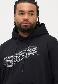 Černá bavlněná mikina s kapucí, na které je bílý grafický potisk s textem a tvary. Kapuce se stahovací šňůrkou a kapsa typu klokanka. Jemná textury.