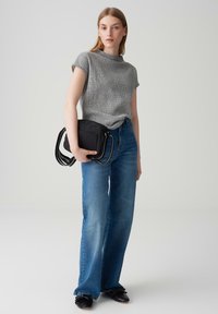 Haut gris texturé à manches courtes, jeans taille haute bleus, sac à bandoulière noir et chaussures noires avec des nœuds. Fond neutre.