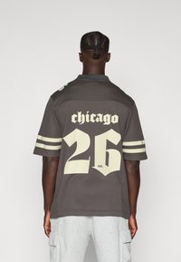 Jerseu gri cu inscripția "Chicago" și numărul "26" în litere mari de culoare crem. Mânecile și gulerul sunt în dungi. Materialul pare texturat.
