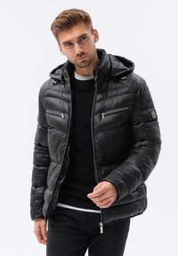 Chaqueta acolchada negra con capucha, con diseño acolchado, bolsillos con cremallera y acentos de herrajes plateados. Lleva puesta sobre un suéter negro.