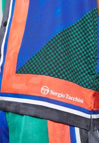 Többszínű ing kék, narancs és zöld részekkel, amelyen kockás minta látható. Fehér és fekete kiegészítők, "Sergio Tacchini" logóval.