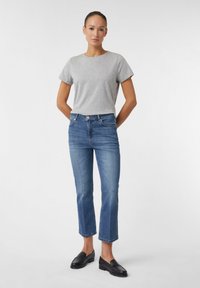 Graues T-Shirt mit kurzen Ärmeln kombiniert mit mittelhochgeschnittenen blauen geraden Jeans. Das Modell trägt schwarze Slipper und präsentiert einen lässigen Look.