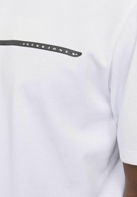 Hvit bomull T-skjorte med en svart horisontal stripe og "JACK & JONES" logo i store bokstaver langs stripen. Korte ermer.
