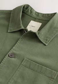 Gros plan sur une chemise boutonnée vert olive avec col et poche poitrine, comportant une étiquette en tissu crème à l'intérieur du col.