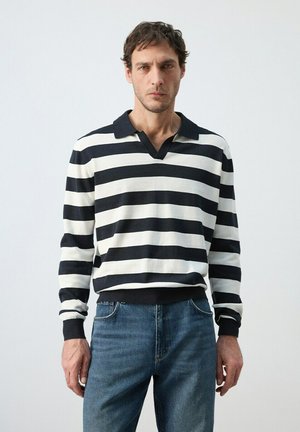 Hombre que lleva una camisa de manga larga a rayas horizontales blancas y negras con cuello y vaqueros azules, de pie frente a un fondo liso y claro.