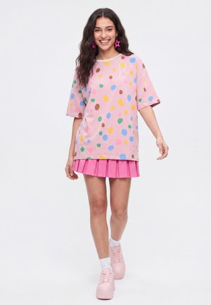 Ung kvinde iført en lyserød oversized polkaprikket t-shirt, lyserød plisseret nederdel, lyserøde sneakers og stjerneformede øreringe går fremad smilende.