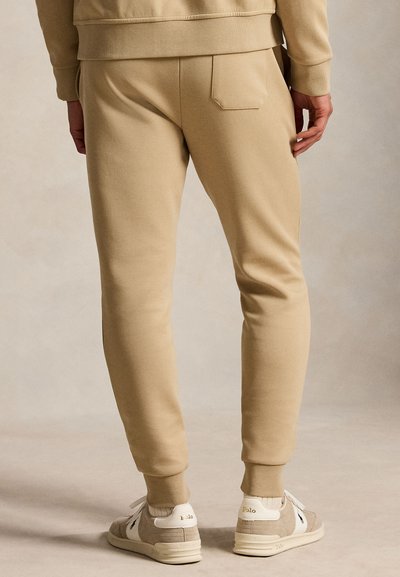 Homme portant un pantalon de survêtement beige, un sweat-shirt beige et des baskets Polo beiges, debout sur un fond neutre, dos à la caméra.