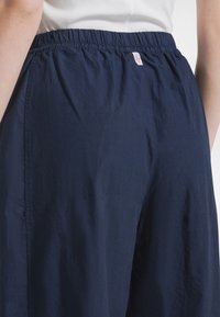 Granatowe spodnie z gumką w pasie o gładkiej, lekkiej fakturze. Posiadają różową metkę z marką w pobliżu pasa. Prosty, luźny krój.