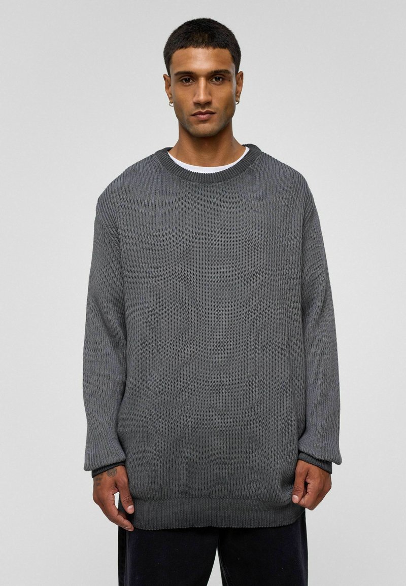 Pull en maille gris avec des manches longues, un col rond et une texture côtelée. Présente une coupe ample et un ourlet épais, porté par-dessus un top blanc.