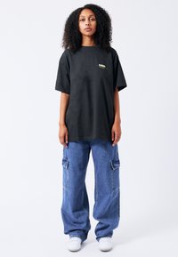 Černé oversized tričko s krátkým rukávem a malým žlutým logem na hrudi, v kombinaci se světle modrými cargo džíny s bočními kapsami.