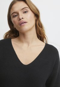 Pull en tricot côtelé noir avec un col en V et des manches larges, présenté avec un design simple et texturé.
