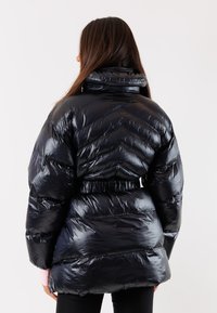 Giacca a vento nera con superficie lucida e trapuntata, caratterizzata da un colletto alto e una vita fasciata; progettata con motivi a chevron.