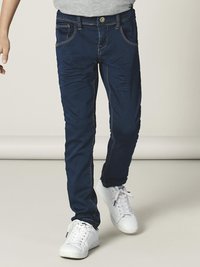 Mørkeblå denimjeans med slim fit, frontlommer med synlige sømmer og knappelukking. Kombinert med hvite joggesko.