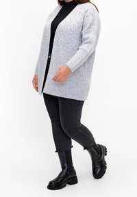 Ljust grått stickat cardigans med ribbad textur, öppen framsdesign och utsmyckade detaljer på axlarna, bärs över en svart topp och jeans.