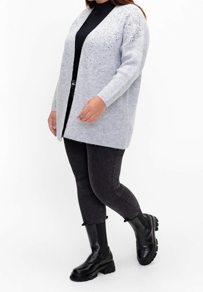 Ljust grått stickat cardigans med ribbad textur, öppen framsdesign och utsmyckade detaljer på axlarna, bärs över en svart topp och jeans.