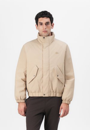 Beige gepolsterte Jacke mit hohem Kragen, elastischen Bündchen, zwei Klapptaschen und taillierter Taille. Mit Logo auf der Brust und glatter Textur.