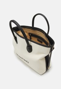 Tygkasse i krämfärg med svarta läderdetaljer och dubbelhandtag, med en beige insida och dragkedjeficka. "VALENTINO" tryckt på framsidan.