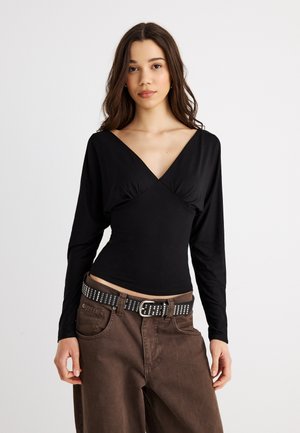 Νέα γυναίκα με μακριά κυματιστά μαλλιά, φορώντας μαύρο τοπ με βαθύ V λαιμόκοψη και μακριά μανίκια, καφέ τζιν και μαύρη ζώνη με τρούκς, στέκεται μπροστά σε λευκό φόντο.