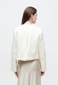 Woman with long brown hair wearing a white textured jacket and beige satin skirt, facing away against a plain white background.