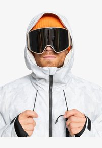 Weiße Camouflage-Ski-Jacke mit hohem Kragen, schwarzen Akzenten und einem Reißverschluss auf der Vorderseite. Dazu gehört eine orange Strickmütze und große spiegelnde Skibrillen.