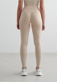 Leggings beige a vita alta con un tessuto liscio e stretch, caratterizzati da un design testurizzato sul retro e abbinati a scarpe da ginnastica bianche.