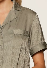 Satinasti pidžama gornji dio u maslinasto zelenoj boji s ponavljajućim uzorkom loga, kratkim rukavima i jednim džepom na prsima. Glatka tekstura, klasični ovratnik.