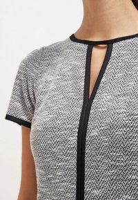 Šedý a bílý top s hřebíčkovým vzorem a černým lemem, s výstřihem ve tvaru klíče a krátkými rukávy. Měkká, strukturovaná látka.