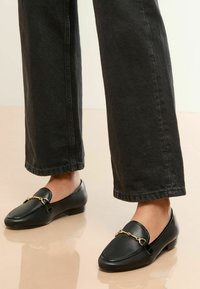 Schwarze Lederloafer mit flachem Absatz, verziert mit einer goldenen Kettenakzent entlang des Vorderfußes und gestickten Details am Rand.