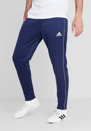Mężczyzna ubrany w granatowe spodnie dresowe Adidas z białym przeszyciem, białe buty Adidas oraz białą koszulę, stojący na tle szarego tła.