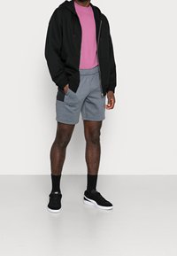 Černá mikina s kapucí na zip, světle růžové tričko, šedé šortky se bočními kapsami a černé tenisky s bílou podrážkou. Neformální sportovní outfit.