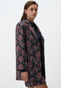 Blazer long et à motifs présentant une base bleu marine sombre avec des motifs floraux complexes en rouge, rose et violet. Coupe ajustée avec une texture lisse.