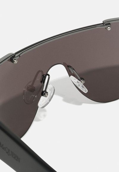 Occhiali da sole neri con lenti scure e curve, dotati di dettagli in metallo e naselli trasparenti. Tempie nero opaco con dettagli di branding.