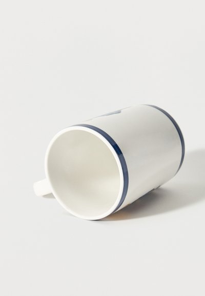 Λευκή κεραμική κούπα με μπλε περίγραμμα, ακουμπισμένη στο πλάι πάνω σε λευκή επιφάνεια, με το χερούλι ορατό στην αριστερή πλευρά.