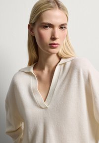 Pull en tricot blanc, doux, avec une coupe décontractée et un décolleté en V, présentant des épaules tombantes et une texture lisse.