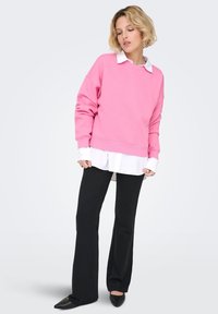 Ροζ φαρδιά φούτερ φορεμένο πάνω από ένα λευκό πουκάμισο με γιακά, συνδυασμένο με μαύρο καμπάνα παντελόνι και μυτερά μαύρα παπούτσια. Λεία υφάσματα.