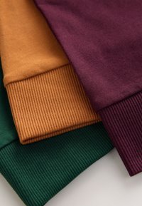 Poignets côtelés pliés empilés de pulls en coton dans des couleurs vert foncé, jaune moutarde et violet profond.