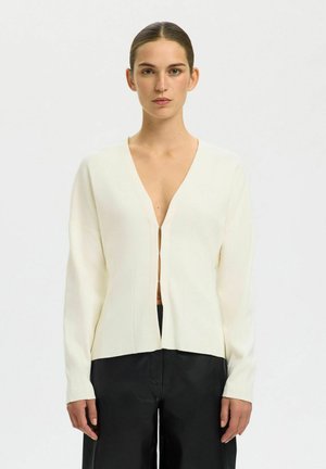 Kobieta z zaczesanymi do tyłu włosami, ubrana w kremowy kardigan z dekoltem w serek zapinany na haczyki oraz czarne spodnie, stojąca na neutralnym tle.