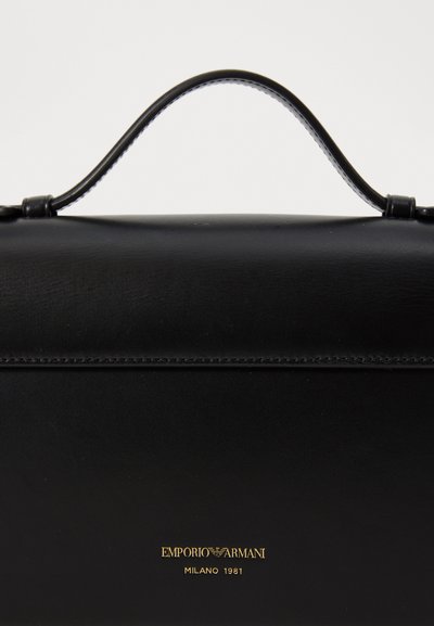 Borsa a mano in pelle nera con manico curvo, caratterizzata da un logo dorato in rilievo e linee pulite e affusolate. Texture liscia con dettagli minimi.