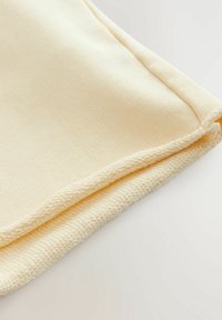 Pehmeä, vaaleankeltainen kangas, jossa on uurrettu rakenne ja sileä reunus. Alahelma on neulottu viimeistelyyn lisättyä yksityiskohtaa varten.