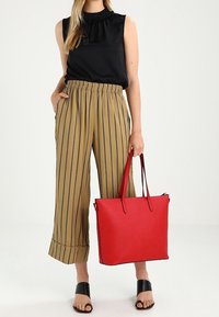 Žena v černém bezramínkovém topu, béžových širokých pruhovaných kalhotách, černých sandálech, držící velkou červenou tašku za ucha.