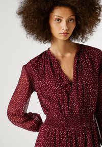 Burgundijas kleita ar plīvētu dizainu, garām caurspīdīgām piedurknēm un smockotu jostu ar baltām punktiņām. Dekoltē ir sakopota.