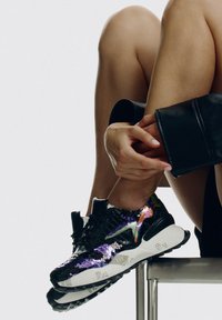 Zapatillas negras con lentejuelas y suela blanca gruesa, acentos multicolores y patrones texturizados. La modelo ajusta una prenda de cuero.