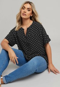 Blusa nera a maniche corte con pois bianchi, dotata di una pattina con bottoni e vestibilità ampia, abbinata a jeans blu. Tessuto liscio.