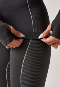 Black athletic leggings with ribbed texture and grey accents. Close-up of hands adjusting the waistband. Features fitted design and long sleeves.