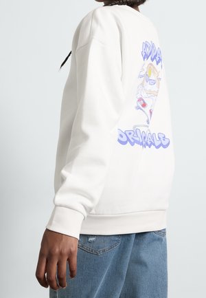 Person iført en hvid sweatshirt med en farverig tegneserie af en skateboardende toast på ryggen og blå jeans.