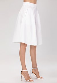 Jupe évasée blanche avec des détails plissés, fabriquée dans un tissu texturé, associée à des sandales à talons hauts blanches.